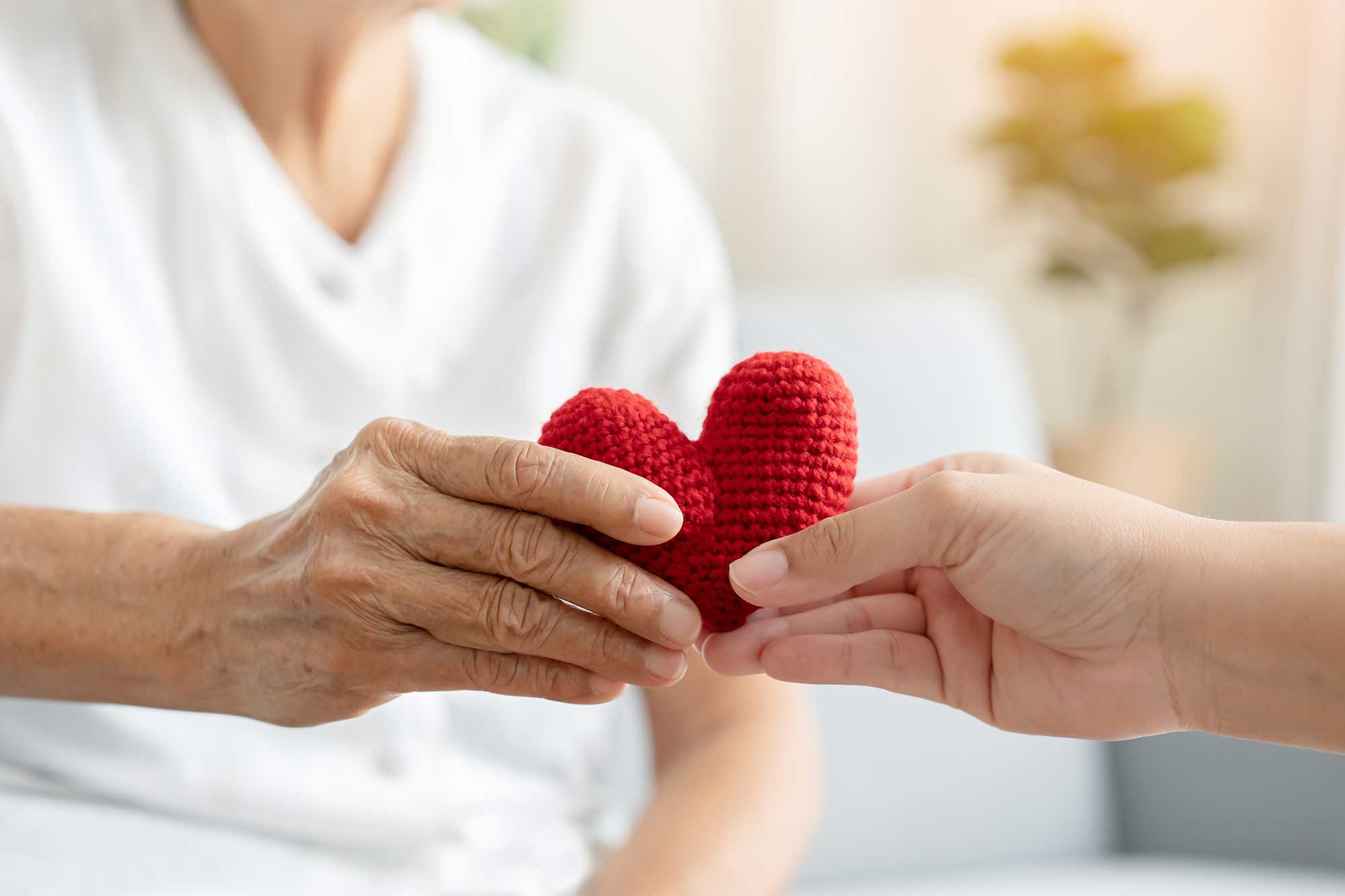 A caregiver in a white uniform gently offers a small crocheted red heart to another person, conveying warmth, compassion, and comfort.