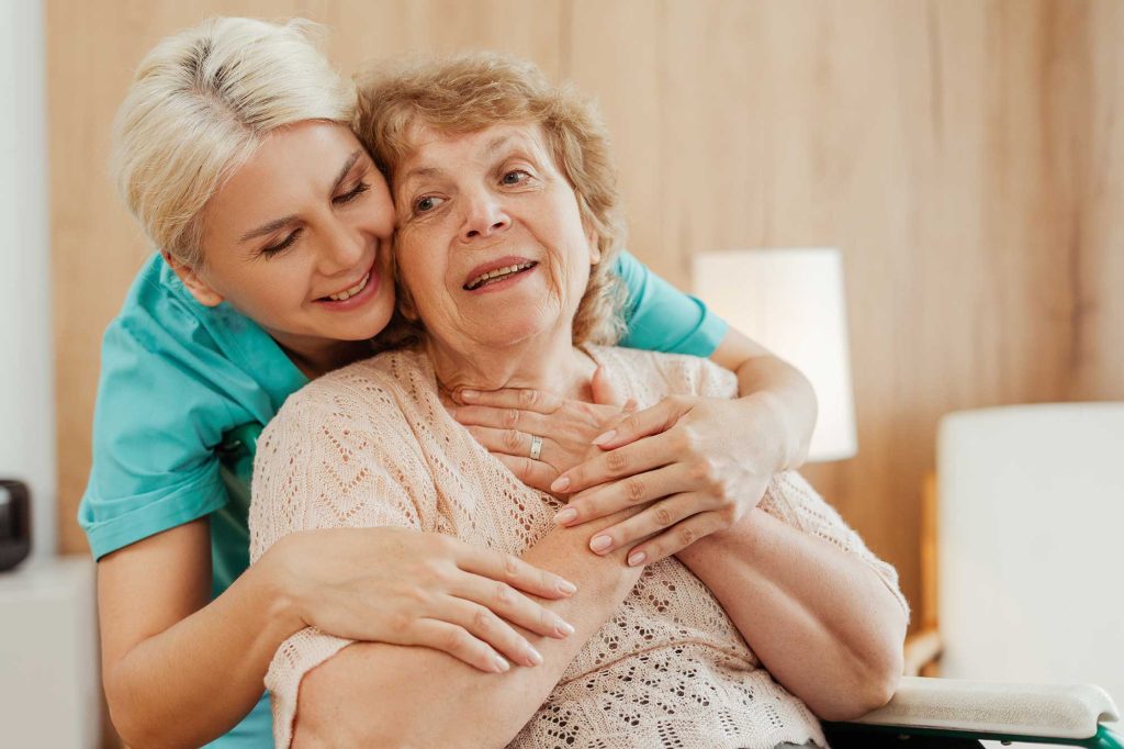 A smiling younger woman in a turquoise shirt hugs an elderly woman in a wheelchair from behind, sharing a tender moment in a warm home.